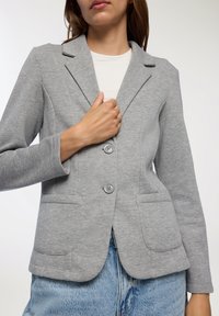 Blazer grigio in jersey con rever a punta, due tasche frontali e chiusura a due bottoni. Presenta una texture liscia e una vestibilità sagomata.