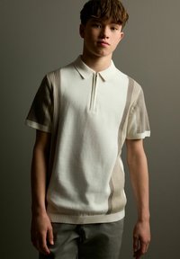 Kortærmet polo skjorte i creme med brune ribbede accentpaneler, der har en krave og en halv-zip fortil. Blød tekstur, minimalistisk design.