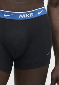 Homme portant un boxer noir Nike avec une ceinture bleue et des logos Nike sur la ceinture et la jambe, debout avec un bras visible sur fond blanc.