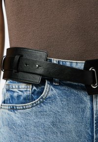 Ceinture en cuir noire au design large, dotée de deux boucles métalliques et d'une texture lisse, portée sur un jean en denim bleu délavé.