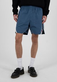 Männlich, trägt blaue Sportshorts mit weißen Seitenstreifen, schwarze Slipper mit Quasten, weiße Sportsocken und einen schwarzen Pullover, steht auf einem weißen Boden.