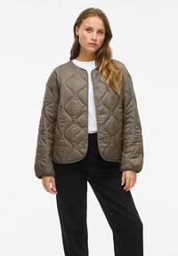 Femme aux longs cheveux portant une veste matelassée taupe à boutons, un chemisier blanc et un pantalon noir, se tenant devant un fond uni.