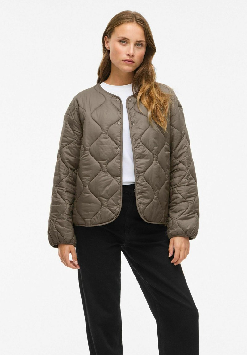 Femme aux longs cheveux portant une veste matelassée taupe à boutons, un chemisier blanc et un pantalon noir, se tenant devant un fond uni.