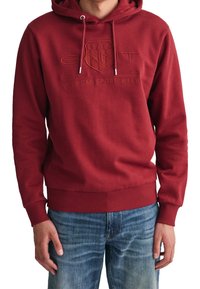 Persoon in een rode hoodie met "GANT 1949 American Sportswear" geborduurd op de borst en blauwe spijkerbroek.