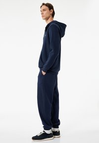 Marineblå hættejakke med frontlomme og justerbar hætte, parret med matchende joggers. Materialet virker blødt og let struktureret. Hvide sneakers.