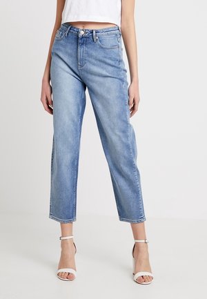 Frau trägt hellblaue Jeans mit hoher Taille, geradem Bein und weißen Sandalen mit Absatz und Knöchelriemen vor einfarbigem Hintergrund.