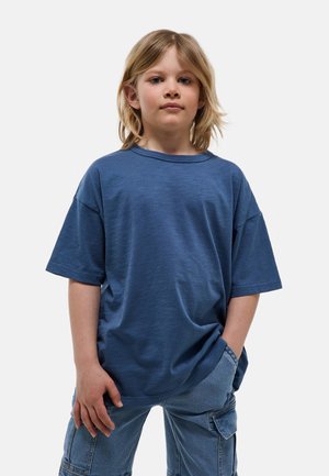 Enfant aux cheveux blonds mi-longs portant un t-shirt bleu ample et un pantalon cargo en denim, debout avec une main dans la poche sur fond blanc.