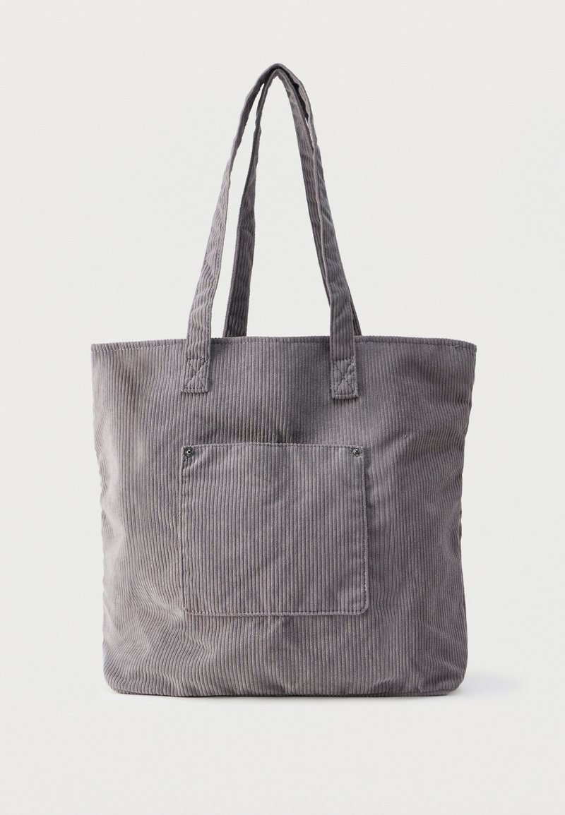 Sac fourre-tout en velours côtelé gris avec de longues anses et une poche plaquée à l'avant, posé sur un fond blanc uni.