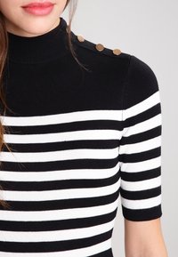 Musta-valkoraidallinen yläosa, jossa on korkea kaulus, lyhyet hihat ja koristeelliset kultaiset napit olkapäällä. Pehmeää neulosta.