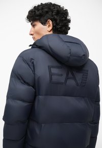 Námornícky modrá puffová bunda s kapucňou, s vyšitým logom "EA" na chrbte, hladká textúra a prešívaný dizajn.