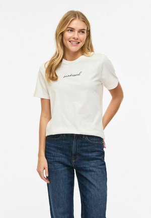 Smilende kvinne med langt blondt hår iført en hvit "sunkissed" t-skjorte og blå jeans, stående med en hånd bak ryggen.