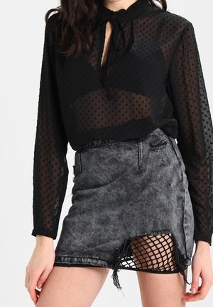 Femme portant un haut noir transparent à pois à manches longues et une jupe en jean gris déchirée avec un ourlet troué laissant apparaître des collants résille noirs.