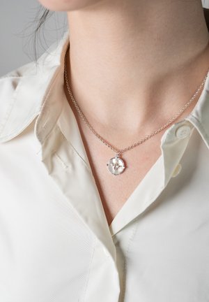 Silverhalsband med en cirkulär medaljong som har en genomskinlig sten, buren över en krämfärgad skjorta med krage.