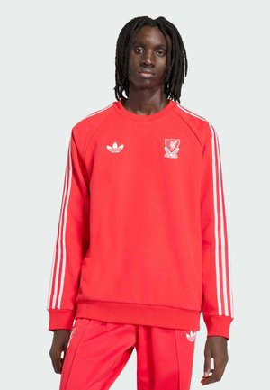 Noor inimene õlgadeni jõudvate dredsidega, kellel on seljas punane Adidas Liverpooli jalgpalliklubi dressipluus heleda halli taustaga.