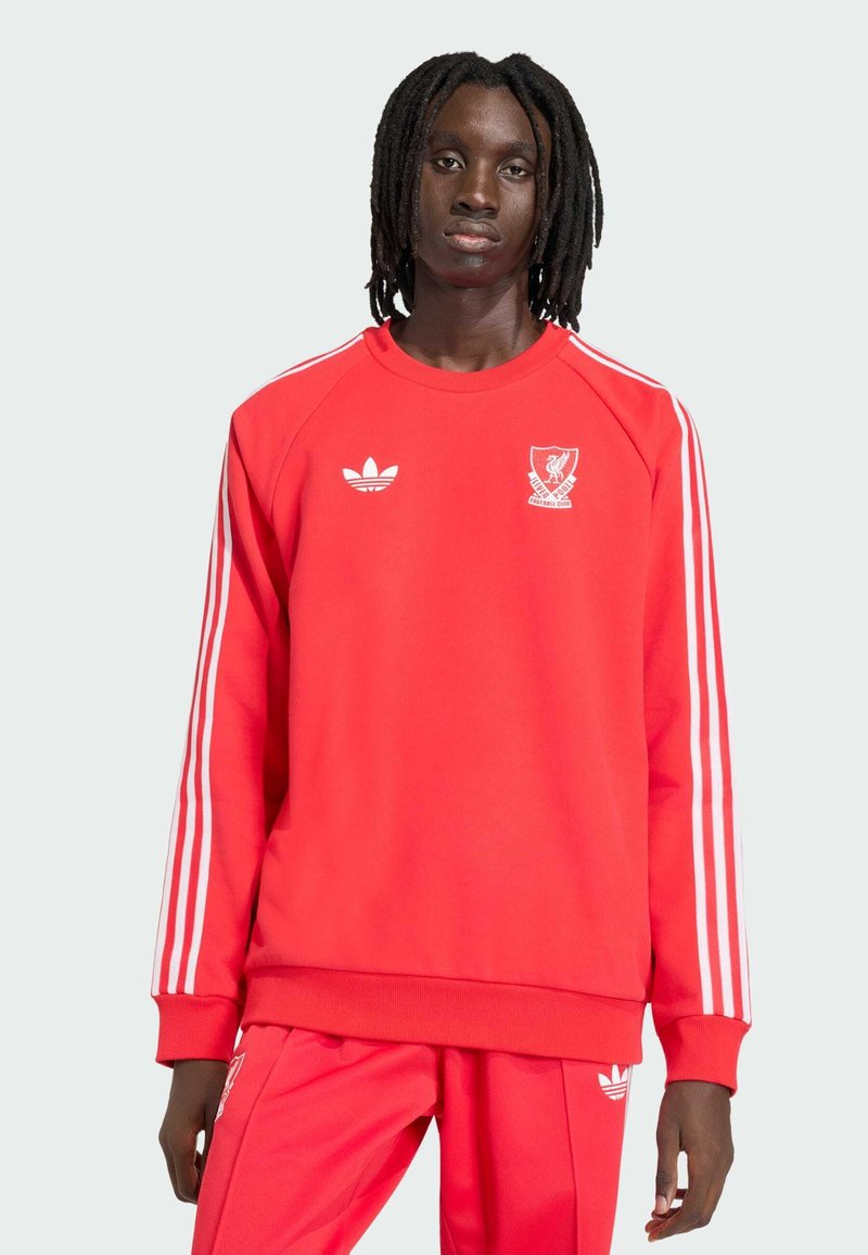 Νεαρό άτομο με dreadlocks έως τον ώμο, φορώντας κόκκινο φόρμας Adidas της Liverpool Football Club, μπροστά από ανοιχτό γκρίζο φόντο.