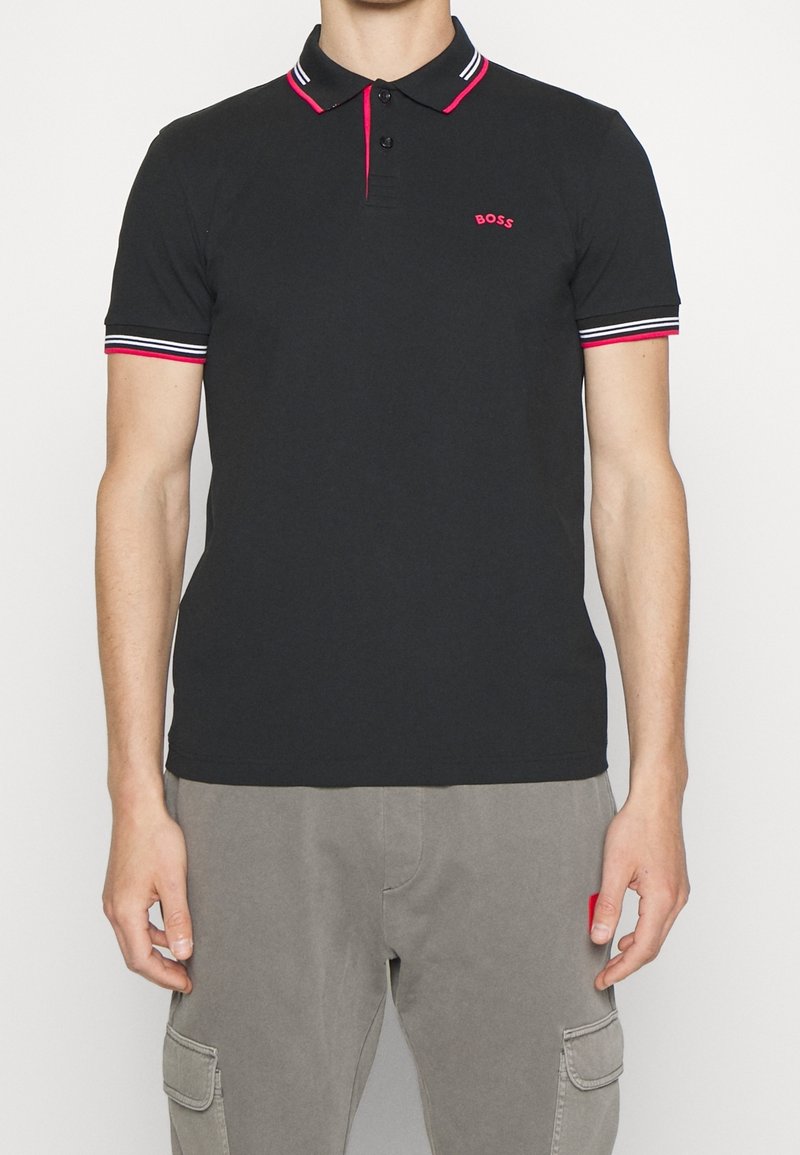 Homme portant un polo noir avec un col et des bords de manches rayés rouge et blanc, petit logo rouge « BOSS » sur la poitrine gauche, et un pantalon cargo gris.