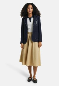 Blazer navy con costuras blancas y emblema, suéter blanco de canalé, falda beige y zapatos negros de slip-on. Texturas suaves, corte ajustado.