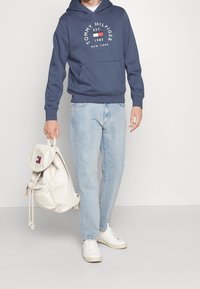 Felpa navy con logo bianco, jeans in denim azzurro chiaro e sneakers bianche. Tiene uno zaino color crema con un cappuccio e cordicella.