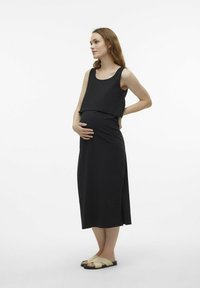 Black maternity dress with a layered design, soft fabric, and ankle length. Worn with light-colored, cross-strap sandals. Plain background.
