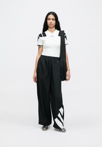 Weißes, eng anliegendes T-Shirt mit schwarzen Schulterakzenten, schwarze locker sitzende Hosen mit großen weißen Streifen und eine schwarze Tasche, die über der Schulter getragen wird.