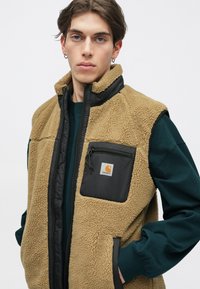 Chaleco de forro polar beige con un panel frontal de nylon negro, bolsillo con cremallera y ribete negro en contraste. Textura suave con un aspecto cálido y acogedor.