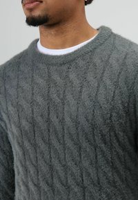 Grijze gebreide trui met een kabelpatroon, ronde hals en een zachte, pluizige textuur. De binnenlaag bestaat uit een wit T-shirt met ronde hals.