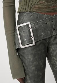 Olivgrön konstläderbälte med en stor silverfärgad rektangulär spänne, med metallöglor längs remmen. Texturerad yta, bred design.