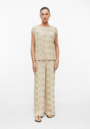Femme debout face à l'objectif, portant un haut ample beige à motifs et un pantalon large, sandales noires, cheveux attachés en arrière, sur fond blanc uni.