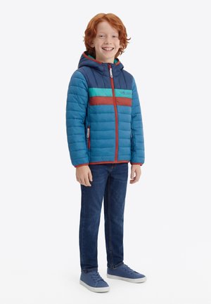 Χαμογελαστό αγόρι με κόκκινα μαλλιά που στέκεται, φορώντας μια μπλε μπουφάν puffer με πράσινες και σκουριασμένες ρίγες, μπλε τζιν και μπλε αθλητικά παπούτσια.