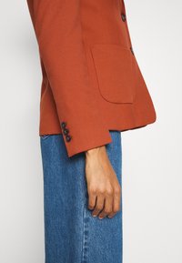 Blazer in roestkleur met een enkele borstzak, met knopen afgewerkte manchetten, gecombineerd met lichtblauwe spijkerbroek. De stof heeft een gladde, gestructureerde afwerking.