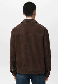 Giacca in suede marrone con colletto, cucitura posteriore singola e polsini con bottoni. Abbinata a jeans blu, la giacca presenta una texture liscia.
