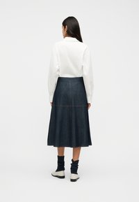 Γυναίκα στέκεται με την πλάτη προς το μέρος μας, φορώντας λευκό πουκάμισο με μακριά μανίκια, σκουρόχρωμη μίντι φούστα από τζιν, σκουρόχρωμες χαλαρές κάλτσες και λευκά παπούτσια με επίπεδη σόλα, μπροστά σε λευκό φόντο.