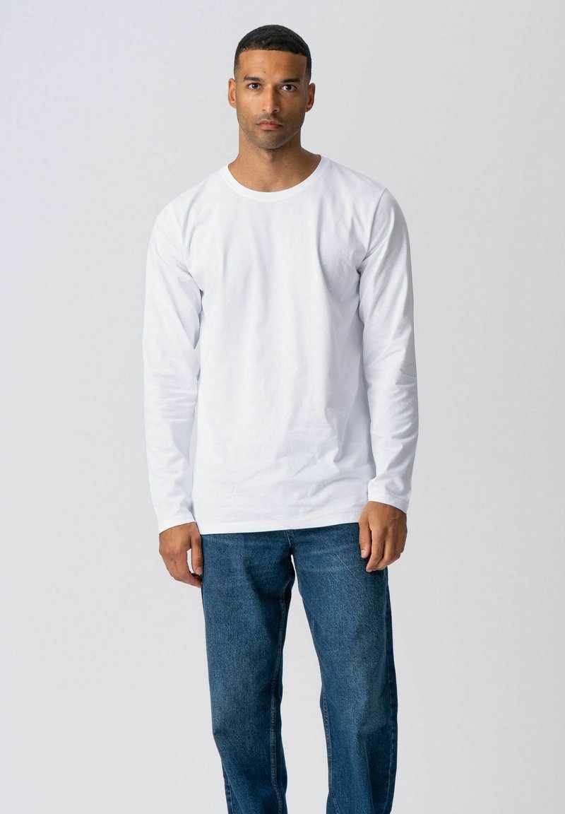 Mand stående med front mod kamera iført en ensfarvet hvid langærmet skjorte og blå jeans mod en neutral baggrund.