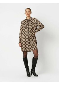 Braun gemustertes Hemdkleid mit Bindebandkragen, das mit Rauten in Creme und Orange verziert ist. Getragen mit kniehohen schwarzen Stiefeln mit Absatz.