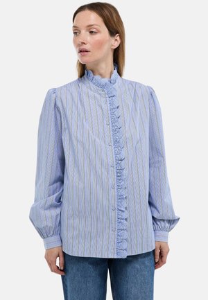 Femme portant un chemisier à rayures bleu clair avec un bord en dentelle le long de la patte de boutonnage et des manches longues bouffantes, associé à un jean bleu.