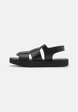 Schwarze Sandale mit flacher Sohle, breiten Riemen und verstellbarem Hinterriemen, gefertigt aus glattem synthetischem Material mit einem minimalistischen Design.