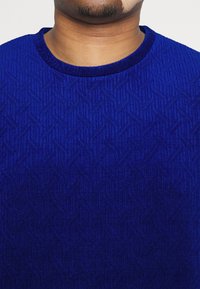 Niebieski sweter w wzory z teksturowym wzornictwem, z okrągłym dekoltem. Tkanina ma gładkie wykończenie i geometryczny wzór.