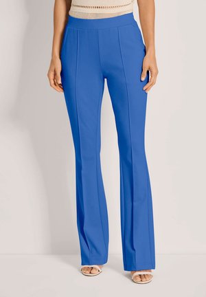 Frau trägt leuchtend blaue, hoch taillierte, ausgestellte Hose, kombiniert mit weißen offenen Sandaletten und einem beigen ärmellosen Top.