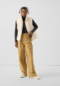 Cremefarbene Fleecweste über einem schwarzen Rollkragenpullover, kombiniert mit beigen Hosen mit weitem Bein und hellen Sneakers; Modell trägt Kopfhörer.