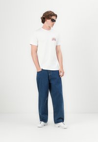 Hvit bomulls t-skjorte med en rød grafikk, kombinert med løstsittende blå denimjeans og hvite sneakers. Solbriller fullfører antrekket.