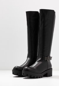 Černé kožené boty po kolena s robustní podrážkou, bočním zipem a detailem popruhu se stříbrnými prvky. Hladká textura a elegantní design.