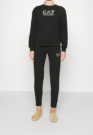 Kobieta stoi w czarnej bluzie i spodniach Emporio Armani EA7, nosi beżowe, puszyste kapcie na gładkim, białym tle.