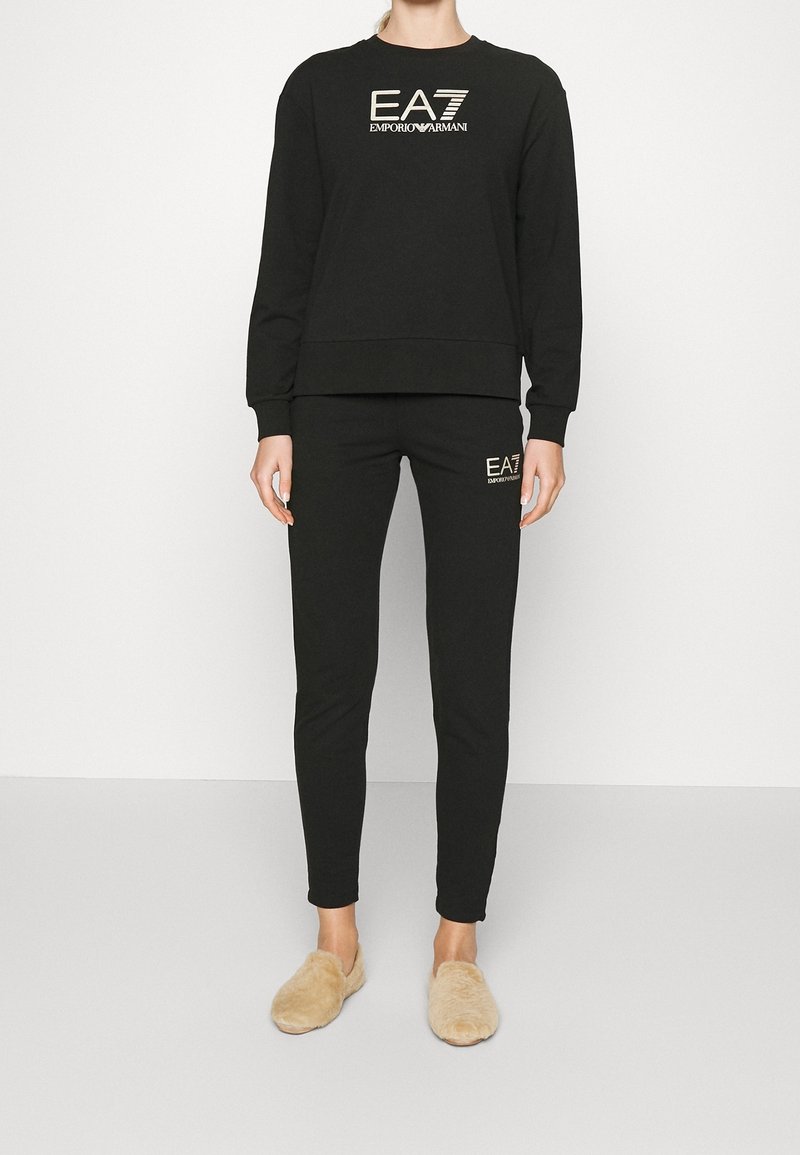 Femme debout en sweat-shirt et pantalon de jogging noirs Emporio Armani EA7, portant des chaussons beiges duveteux sur un fond blanc uni.