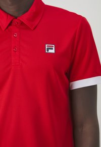 Polo rouge en tissu texturé, doté d'un col et de trois boutons. Il présente une touche blanche sur les manches et un logo carré sur la poitrine.