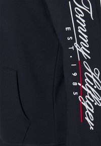 Manga de sudadera azul marino con texto en cursiva blanco "Tommy Hilfiger", "EST. 1985" y un detalle de línea roja vertical.