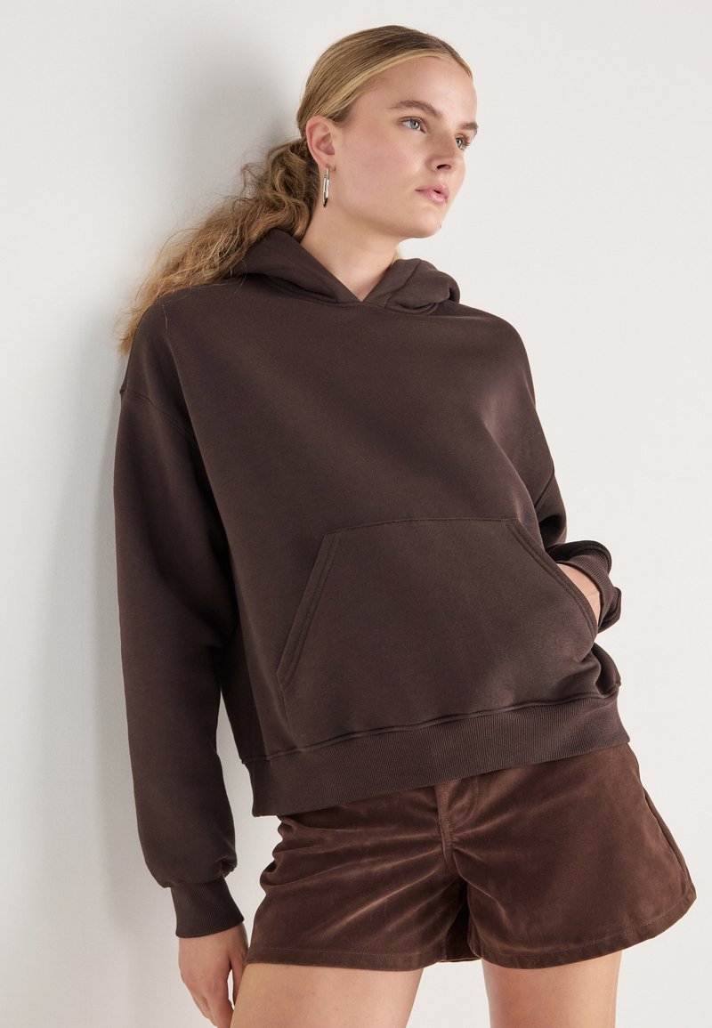 Hanorac maro cu buzunar frontal, croială lejeră și mansete ribelate. Asortat cu pantaloni scurți din suede maro, textură netedă și design minimalist.