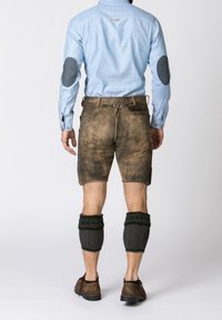 Muž oblečený v světle modré košili s náplety na loktech, hnědých kožených šortkách, tmavých pletených teplých ponožkách a hnědých botách, stojící zády.