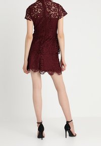 Burgundy spetsklänning med korta ärmar, vågformad nederkant och intrikata blommönster, bärs med svarta högklackade sandaler.