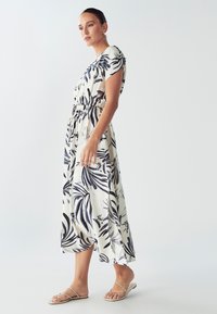 Maxi obleka iz lahkega materiala z belo podlago in mornarskimi listi, ki ima kratke rokave in elastičen pas. Nosena z ravnimi sandali.