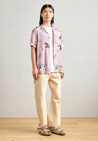 Kortermet rosa mønstrete skjorte med svarte og røde detaljer, kombinert med beige vide bukser og grå sandaler på et treverk gulv.